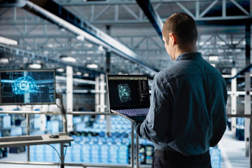 Tecnico con laptop analizza dati AI in una smart factory con magazzino automatizzato sullo sfondo