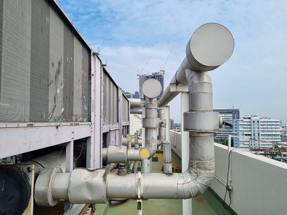 Tubazioni e componenti di un impianto di refrigerazione industriale installati sul tetto di un edificio, con torri di raffreddamento sullo sfondo.